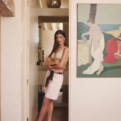 Mulher com top branco de renda e saia branca, com lenço na cintura, em sala com parede branca e pintura colorida