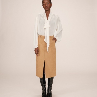 Conjunto feminino com blusa branca de tecido leve e saia camel midi com fenda, botas pretas altas.