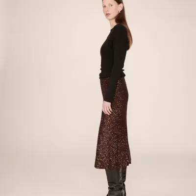 Mulher de perfil com camisa preta, saia castanha brilhante e botas pretas.