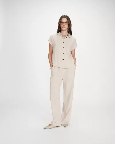 Mulher vestindo conjunto bege claro com camisa de manga curta e calças compridas, sapatos baixos bege e acessórios dourados em fundo branco.