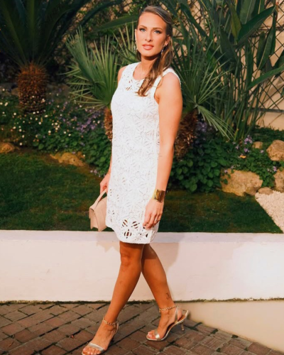 Mulher com vestido branco de renda e sandálias douradas num jardim.