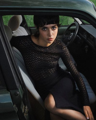 Mulher sentada no banco dianteiro de um carro com blusa preta transparente e saia preta.