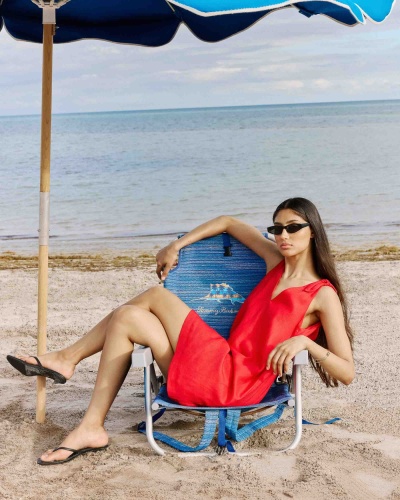 Mulher sentada numa cadeira de praia azul, vestindo vestido vermelho e óculos de sol pretos, na areia junto ao mar