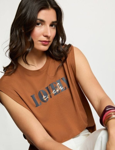 T-shirt castanha sem mangas com texto LOVELY fresh usada por mulher com pulseiras, fundo branco.
