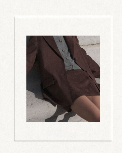 Blazer e saia castanhos com camisola cinzenta e botões, sentada junto a uma parede clara.