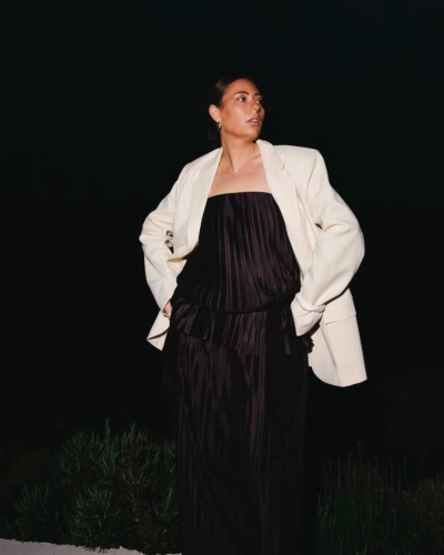 Pessoa com vestido preto plissado e casaco branco num ambiente noturno