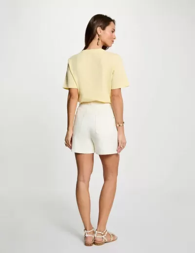 Mulher de costas com t-shirt amarela, calções brancos e sandálias brancas com tachas