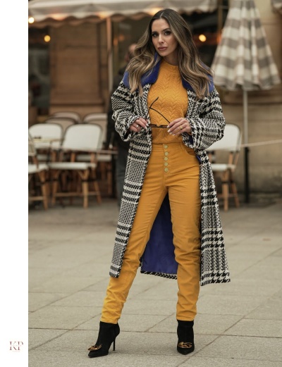 Mulher com casaco pied-de-poule, camisola e calças amarelo mostarda, botins pretos, segura óculos, ao ar livre