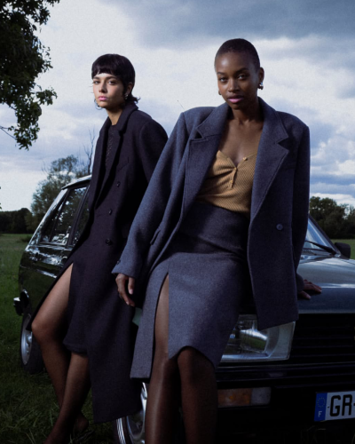 Duas mulheres com casacos compridos e fendas laterais apoiadas num carro antigo preto ao ar livre.