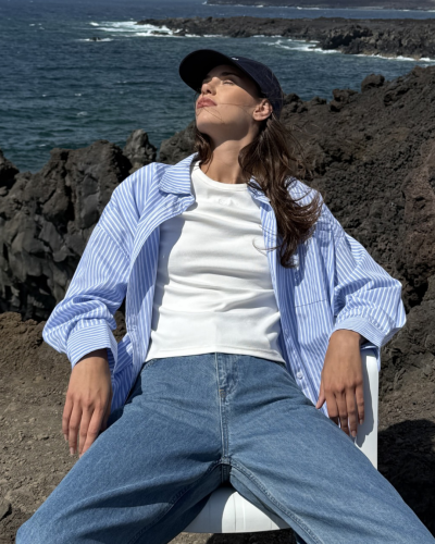 Mulher sentada ao ar livre com camisa azul às riscas, t-shirt branca, jeans e boné preto.