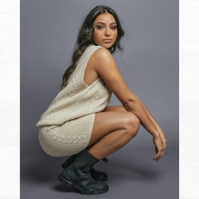 Mulher com conjunto de malha bege e botas pretas num fundo cinza