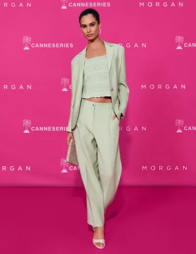 Modelo veste conjunto verde claro com blazer, calças e top de renda, segura mala brilhante, em fundo rosa com textos.