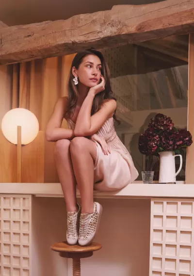 Mulher com vestido rosa claro e sapatilhas bege com detalhes dourados, sentada numa bancada branca em ambiente interior com madeira e cortinas.