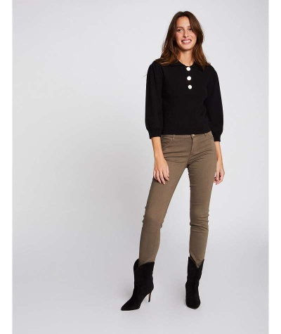 Mulher vestindo camisola preta com botões brancos e calças castanhas, botas pretas, em fundo cinzento claro