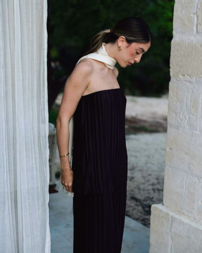 Mulher vestindo conjunto preto plissado e lenço branco ao pescoço em ambiente externo com parede de pedra e cortina branca.