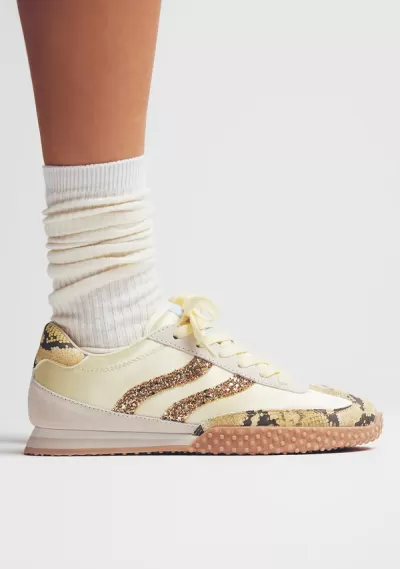 Ténis creme com detalhes em pele de cobra e brilho, solo de borracha texturada, usado com meias brancas.