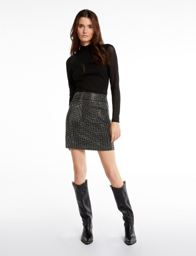 Mulher vestindo saia curta cinza com padrão pied-de-poule e camiseta preta com mangas compridas, usando botas pretas até ao joelho.