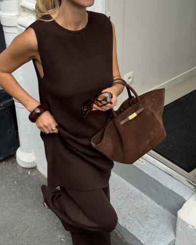 Mulher com vestido castanho escuro segurando mala castanha de camurça e óculos de sol.