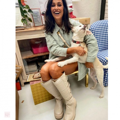 Mulher sorridente com casaco cinzento e botas bege segurando cão branco em sala