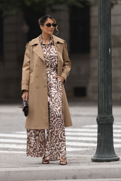 Mulher com casaco bege e vestido com padrão castanho e branco numa rua urbana