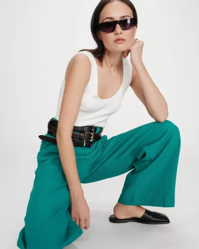 Mulher com óculos de sol, blusa branca, calças verdes largas, cinto preto e sapatos pretos num fundo branco