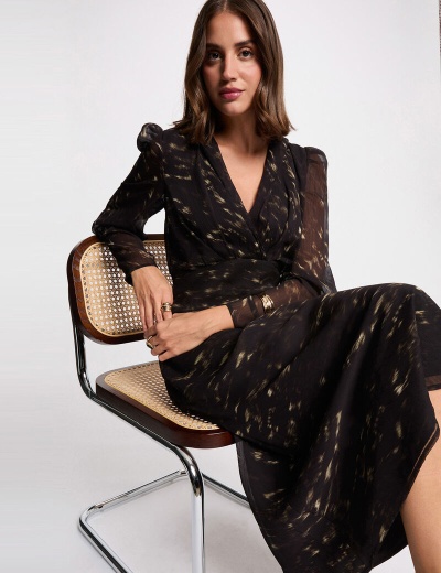 Vestido preto comprido com padrão dourado e mangas transparentes, mulher sentada numa cadeira palhinha e metal.