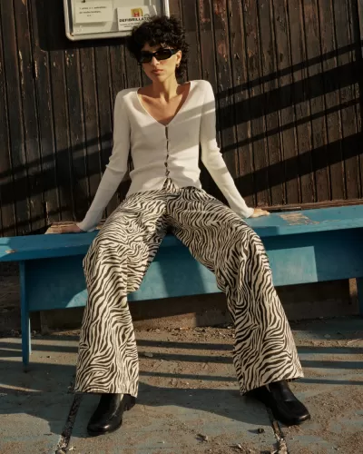 Mulher com sweater branco, calças de padrão animal preto e branco, botas pretas e óculos escuros sentada num banco azul