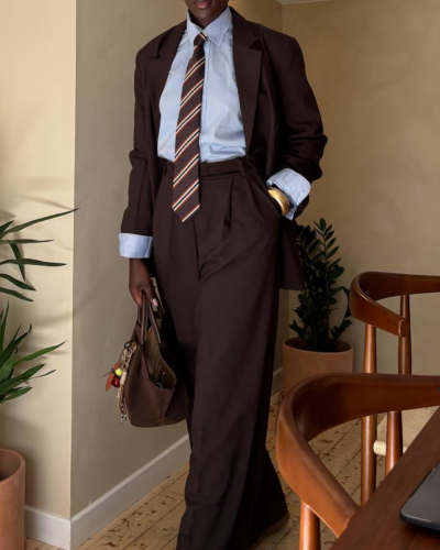 Pessoa com fato castanho escuro, camisa azul, gravata riscada e mala castanha em ambiente interior com plantas.