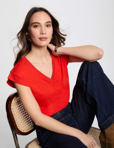 Top vermelho de malha com decote em V e calças de ganga azul escuras sentado numa cadeira com ráfia