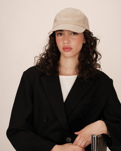 Mulher com casaco preto, camiseta branca e boné bege com texto branco no fundo neutro