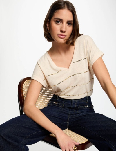 Mulher com camisola creme à riscas douradas e jeans escuros sentada numa cadeira de madeira com palhinha.