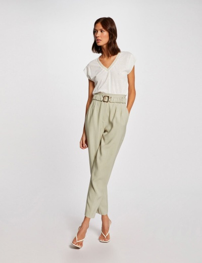 Mulher com t-shirt branca de gola V, calças bege com cinto largo e sapatos brancos de salto, fundo branco.