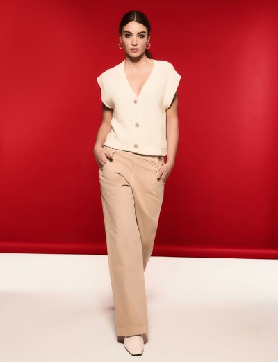 Mulher com colete creme de malha e calças largas bege num fundo vermelho