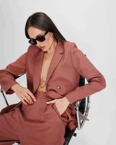 Mulher com fato rosa velho, óculos escuros, sentada numa cadeira de escritório com fundo branco.