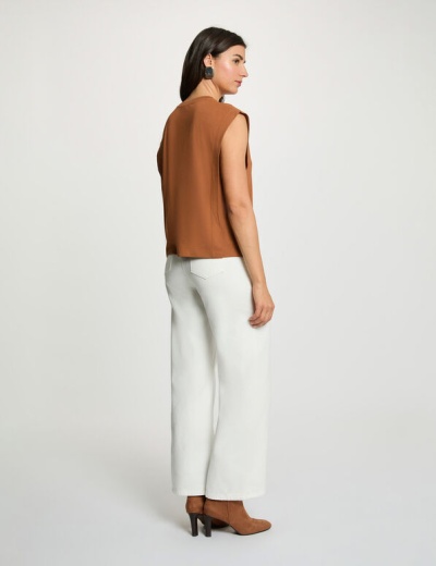 Mulher de costas com colete castanho, calças brancas largas e botas castanhas de salto.