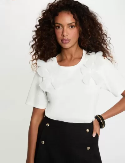 Mulher com t-shirt branca com detalhes em correntes nos ombros e saia preta com botões dourados