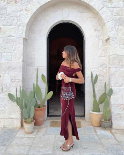 Mulher com vestido bordô longo e saco bordado à frente de porta arqueada de pedra com vasos de cactos.