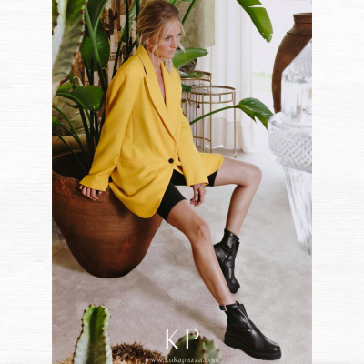 Mulher sentada com blazer amarelo, calções pretos e botins pretos em ambiente interior