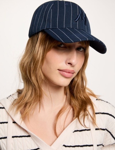 Boné azul escuro com riscas brancas e logótipo, usado por mulher com casaco riscado