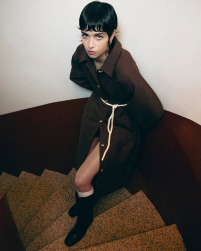 Mulher de casaco castanho escuro com cinto branco numa escada interior