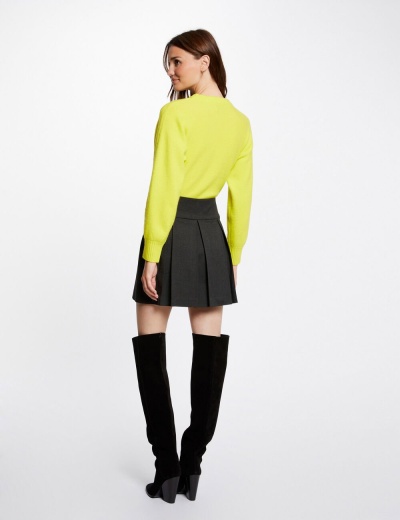Mulher de costas com camisola amarela, saia preta plissada e botas pretas de salto em fundo branco