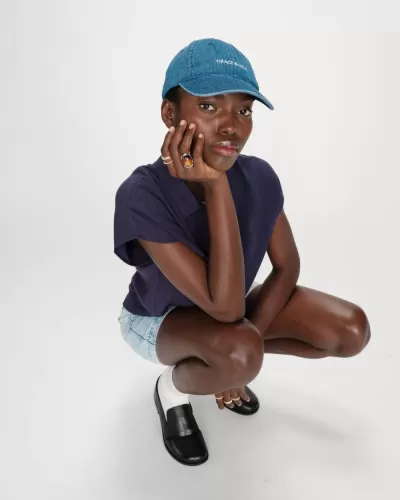 Pessoa com boné azul, camisa roxa, calções de ganga e sapatos negros agachada sobre fundo branco