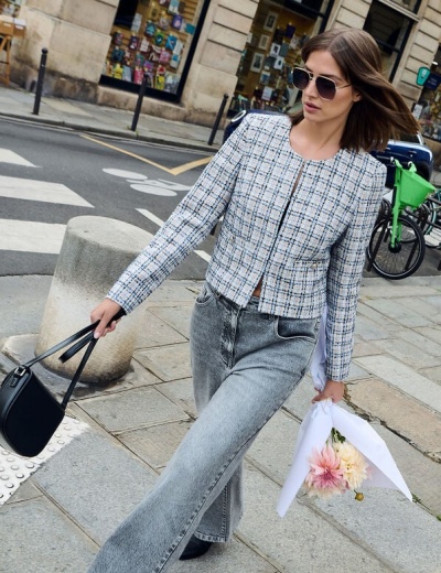 Mulher com casaco tweed azul e branco, jeans cinza, óculos escuros, mala preta e bouquet de flores numa rua urbana