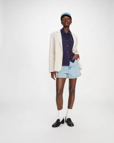 Mulher com blazer bege, camisa azul escura, calções de ganga, sapatos pretos e boné azul