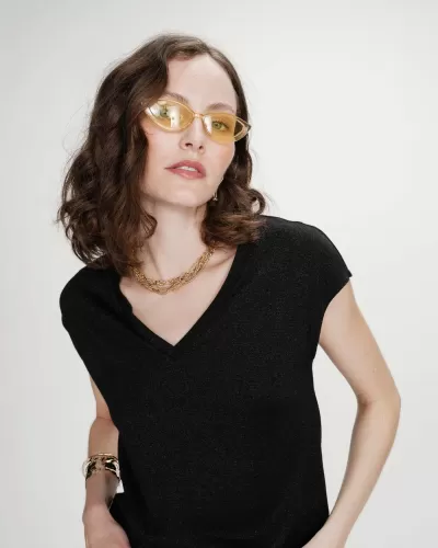 Mulher de cabelo castanho ondulado com camisa preta, óculos amarelos, colar e pulseira dourados, fundo branco.