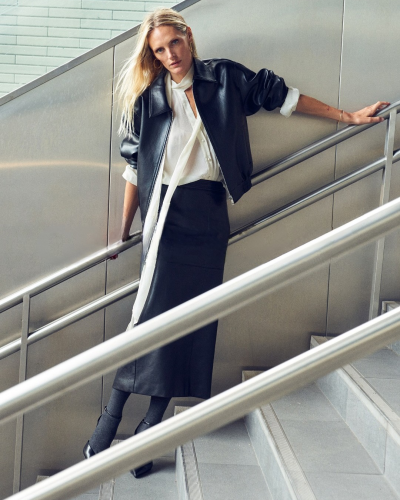 Mulher vestindo casaco de couro preto, blusa branca e saia preta numa escada.
