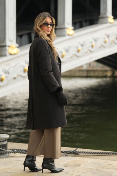 Mulher com sobretudo preto, calças castanhas e botas pretas junto a ponte dourada