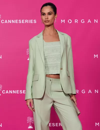 Fato bege claro de mulher com blazer, calças e top recortado, mala prateada, em fundo rosa com texto CANNESERIES e MORGAN.