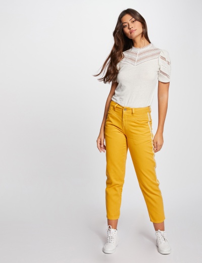 Mulher com calças mostarda até ao tornozelo, t-shirt branca com renda, ténis brancos, fundo neutro