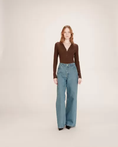 Mulher vestida com camisola castanha de malha, calças de ganga largas azuis e sapatos pretos com fundo claro.